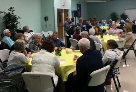 People seated at round tables in a community hall, chatting during a social gathering.