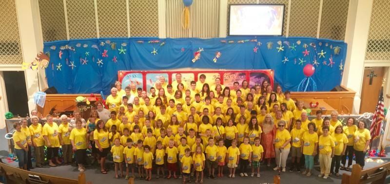 Large group in yellow shirts posing on a stage with a colorful blue backdrop and red arch