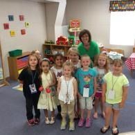 Teacher with a group of children standing in a classroom, smiling at the camera.