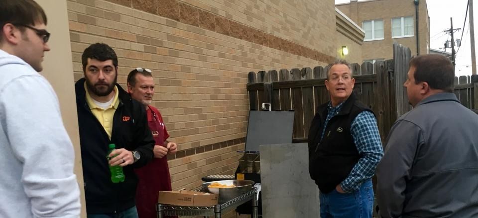 Several people stand and chat beside a brick wall and barbecue grill under a covered outdoor area.