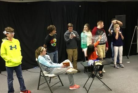 Youth group of people standing and sitting on a stage in front of a black curtain, one person filming on a phone.