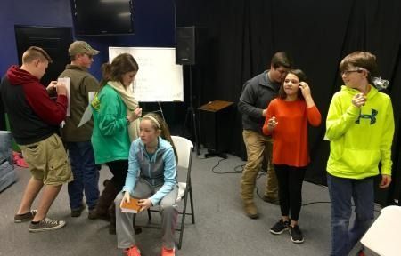 Several people stand and sit in a room, with one seated girl holding a paper and others talking nearby.