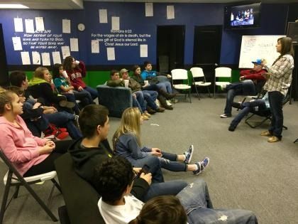 People seated in a classroom circle, listening to a speaker at the front.