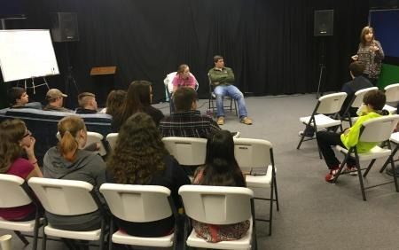 People seated in chairs facing two speakers on a stage in a small auditorium.