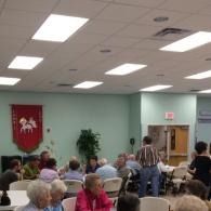 Community meeting in a hall with seated attendees, ceiling lights, and a red banner on the wall