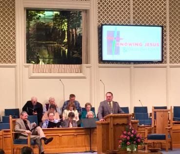 Church service with speaker at pulpit, choir seated behind, and slide reading “KNOWING JESUS” on screen.