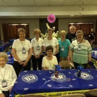 Group posing behind a blue tablecloth-covered table in a hall, with purple decorations and event badges.