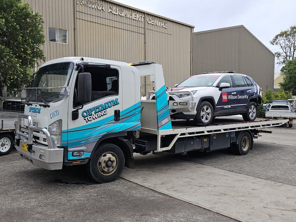 A Tow Truck With a Car on the Back is Parked in Front of a Building — Optimum Towing in Sanctuary Point, NSW