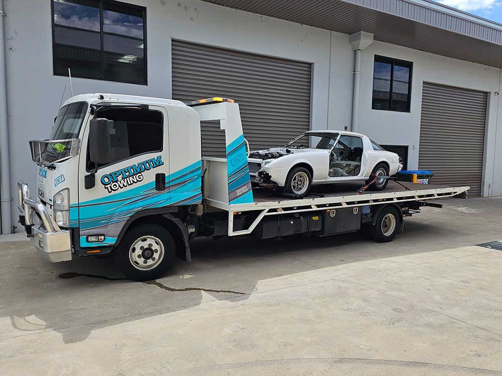 A Tow Truck With A White Car On The Back Is Parked In Front Of A Building — Optimum Towing in Bomaderry, NSW