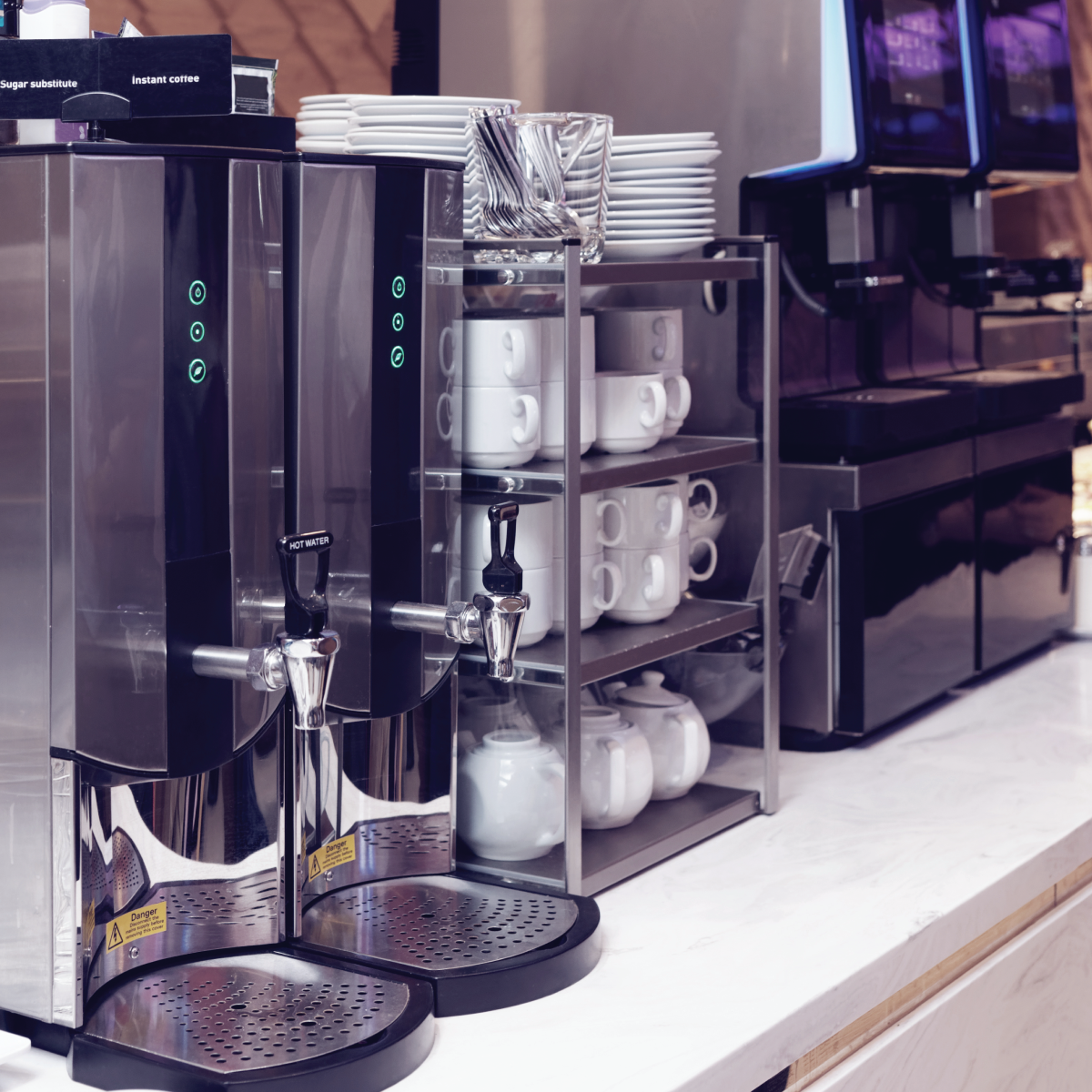 Coffee station with two stainless steel espresso machines and stacked white mugs on shelves.