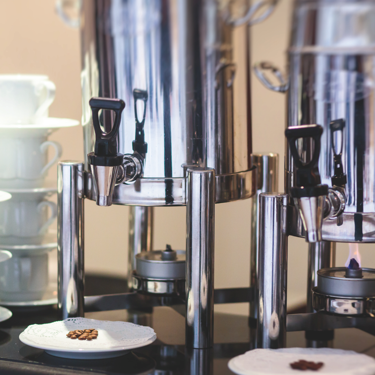 Chrome buffet coffee urns with black spigots beside a plate of coffee beans on a counter