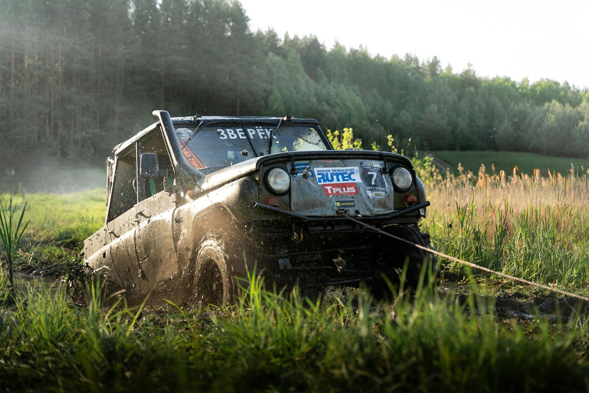Off-road vehicle stuck in muddy field, being pulled by a winch, water splashing.