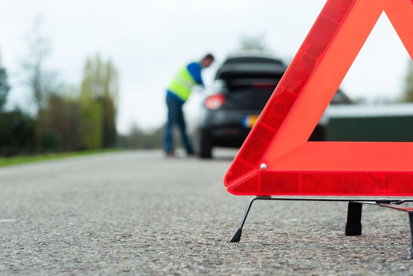 Red reflective warning triangle on roadside, man near disabled car with opened trunk.