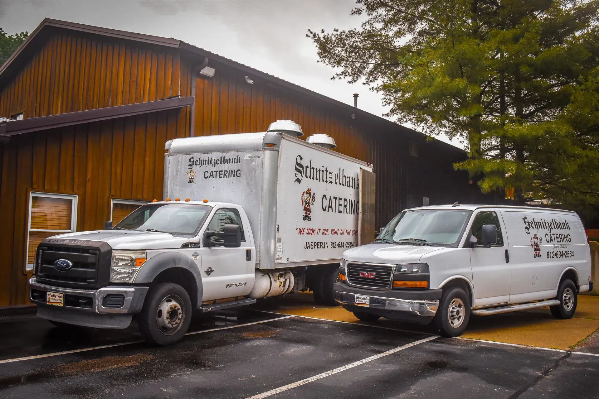 Two white delivery trucks parked beside a wooden building on a wet street