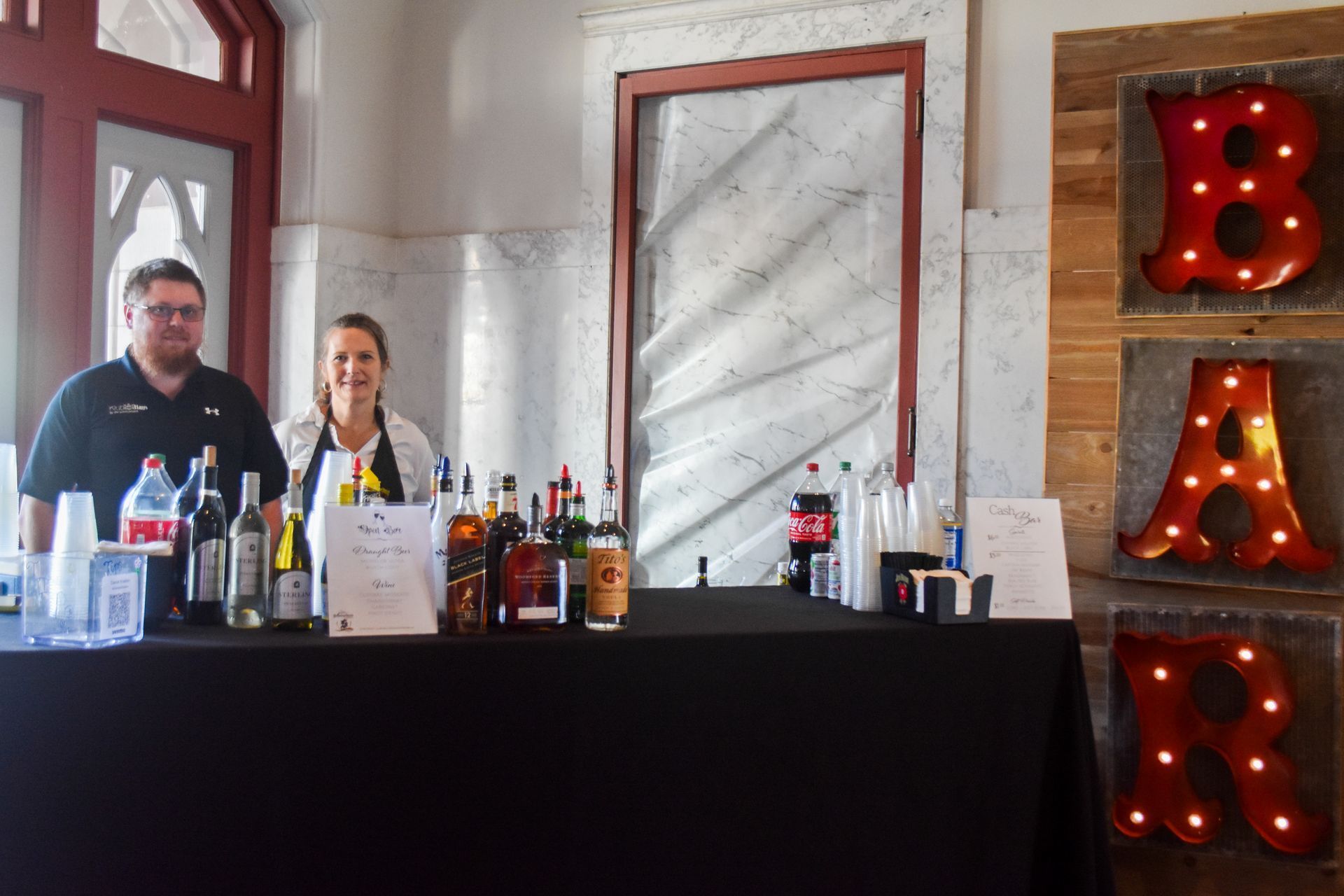 Two people behind a bar table with bottles, beside a lit red “BAR” sign.