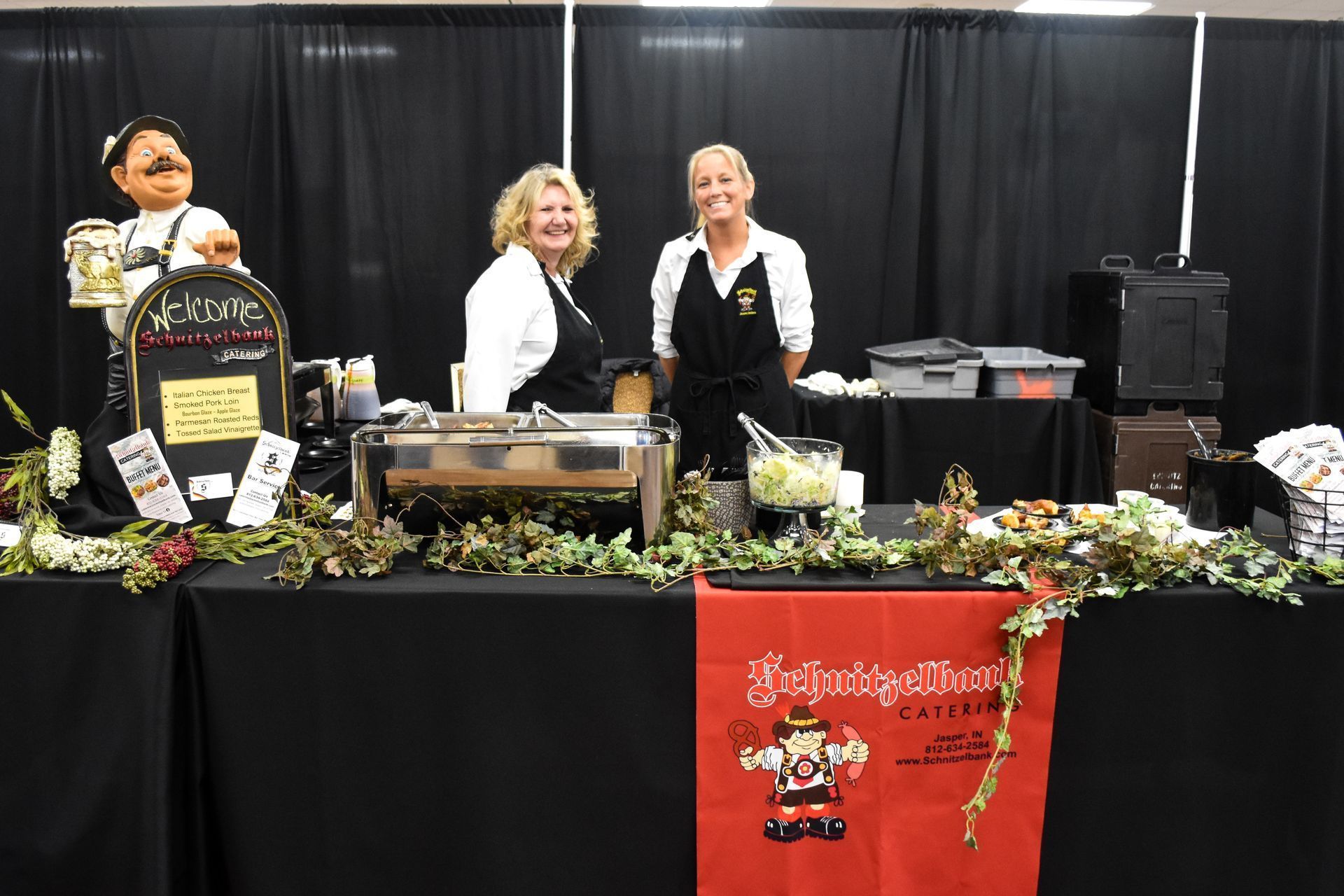 Three people at a booth with plants, signs, and a red table banner in front of a black backdrop.