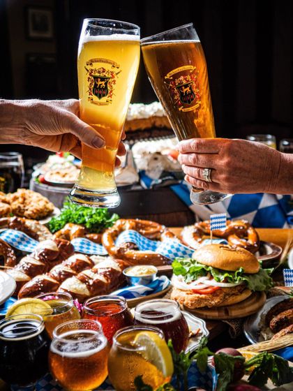 Hands clinking beer glasses over a table of pretzels, burgers, and dips in a cozy restaurant setting