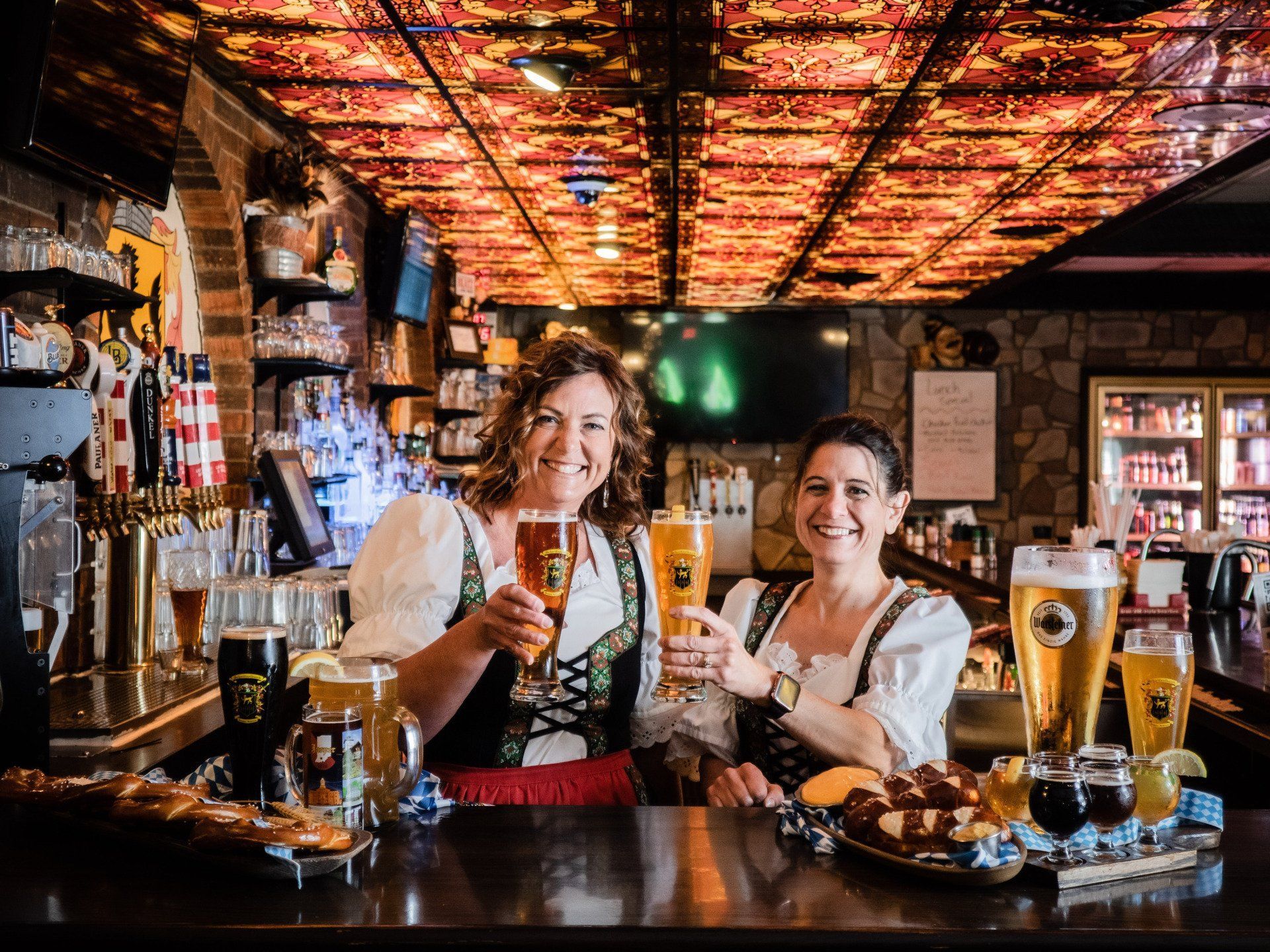 Two bartenders smiling and raising beer mugs behind a bar with warm orange ceiling lights.