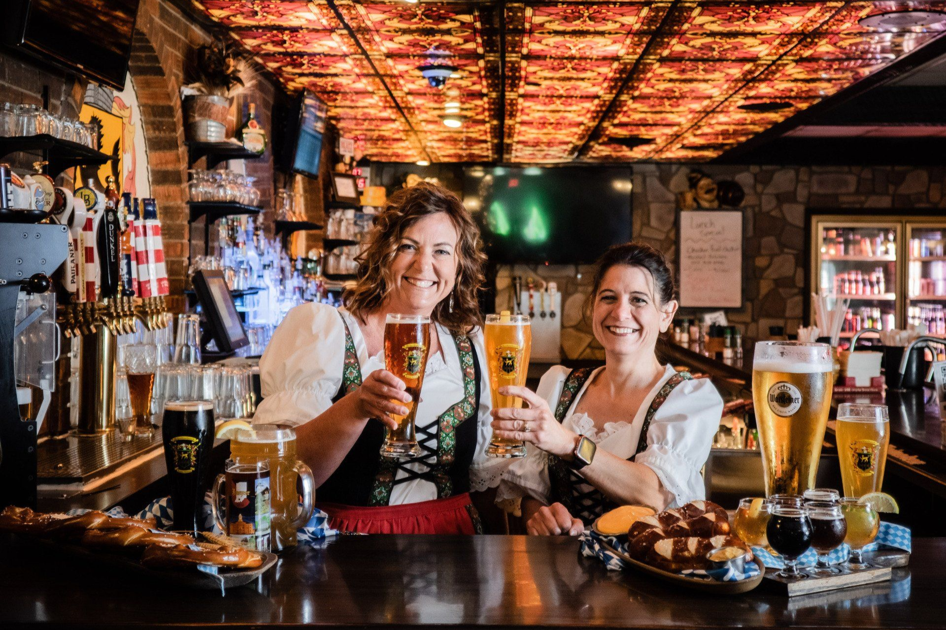 Two bartenders smiling behind a bar, holding pints in a cozy, warmly lit pub.