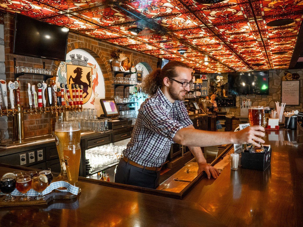 Bartender pouring a drink behind a wood bar in a warmly lit pub with a patterned ceiling.