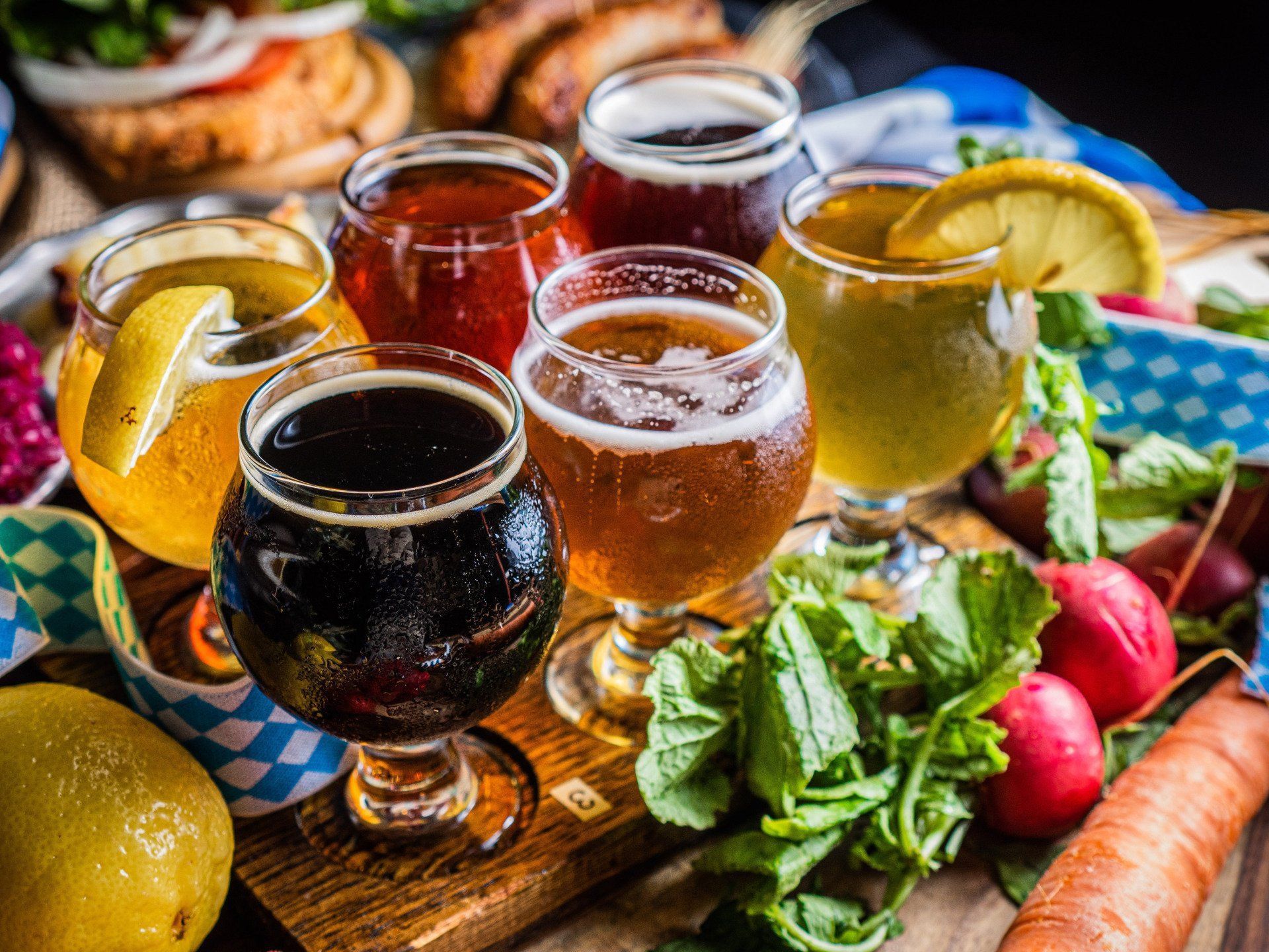 Four glasses of craft beer on a picnic table with lemons, radishes, greens, and a carrot