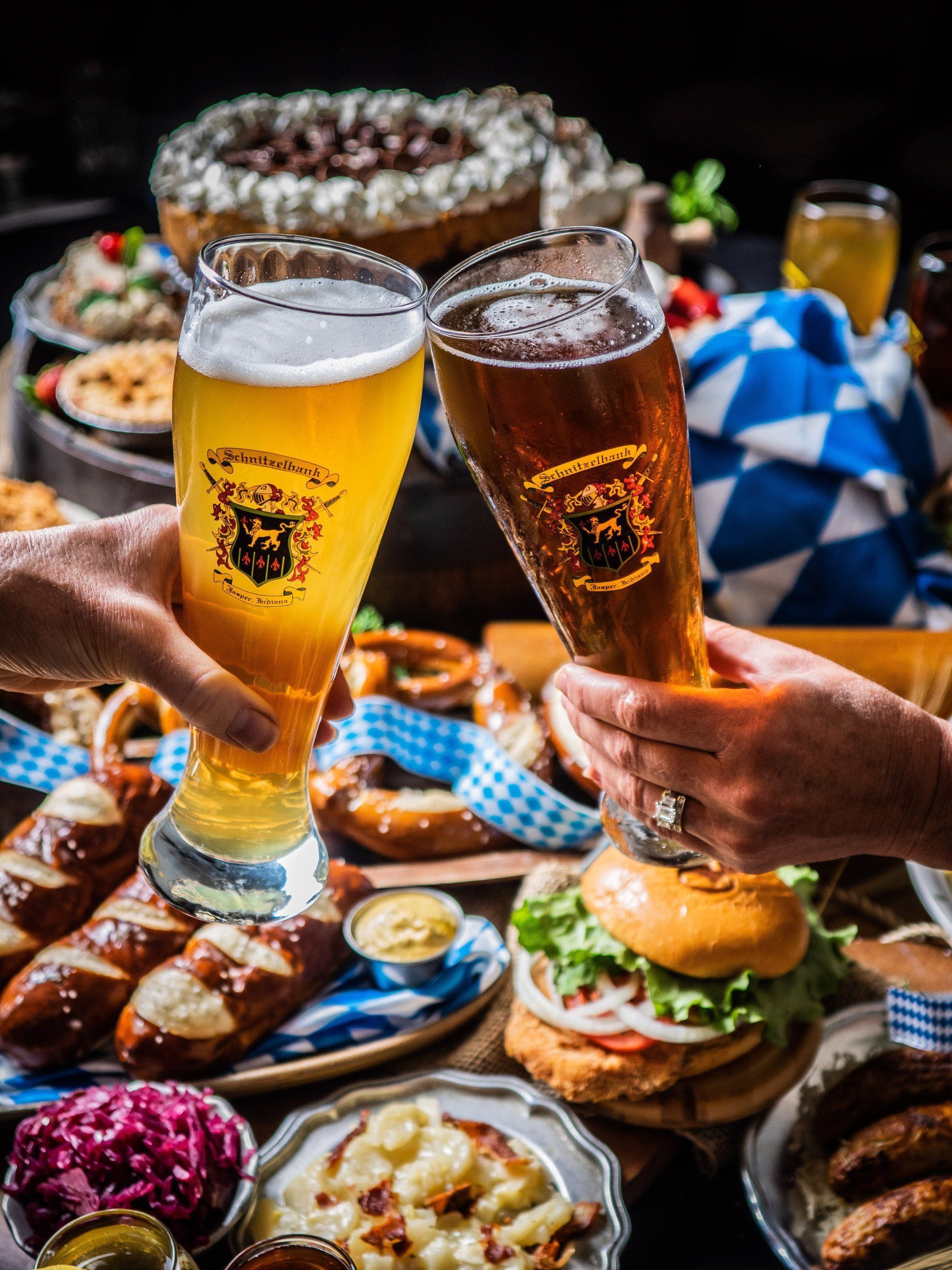 Two hands clink beer glasses over a table of burgers, fries, and Bavarian snacks in a lively setting
