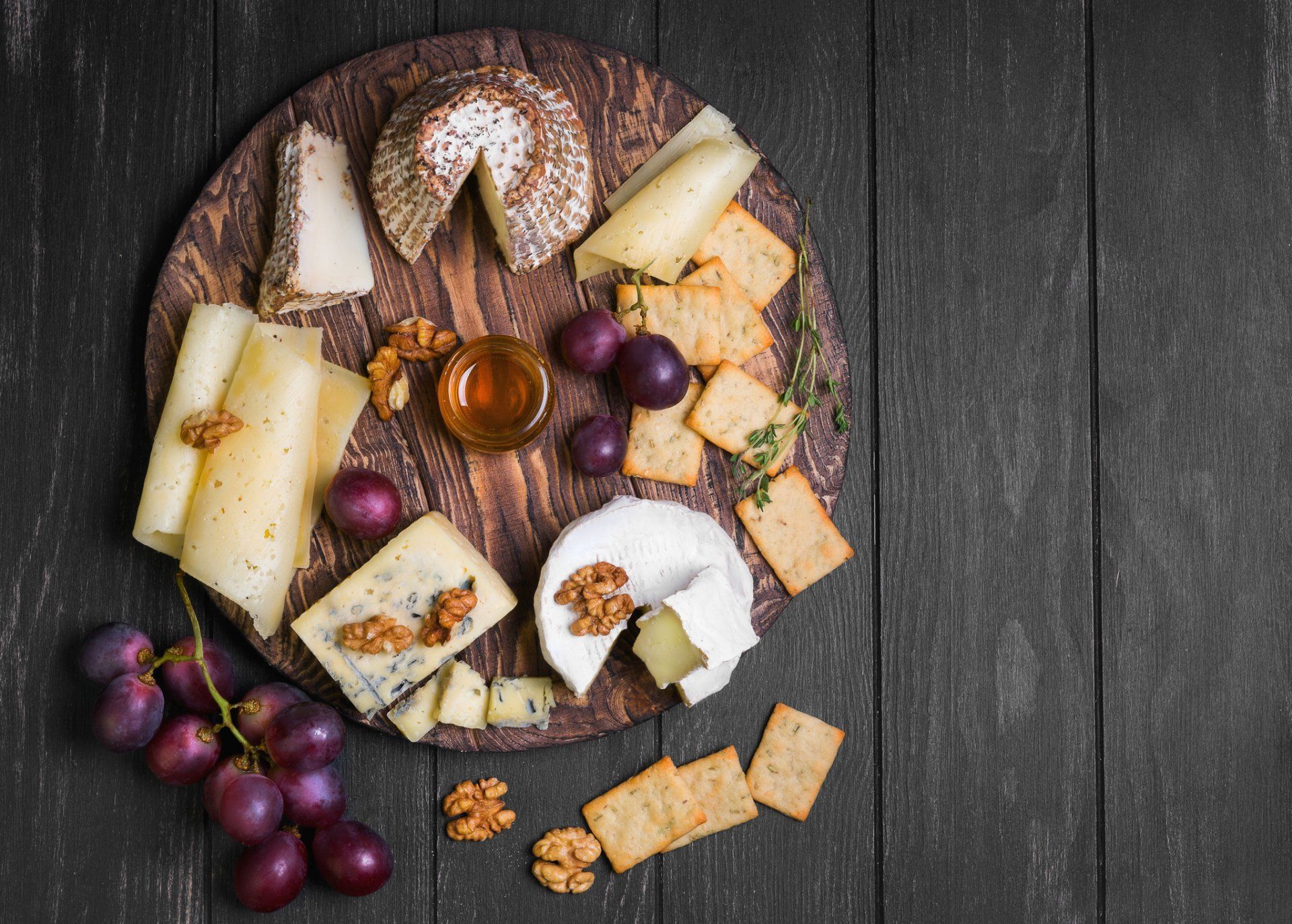 Cheese and grapes on a wooden board with crackers, nuts, and a small bowl of dip