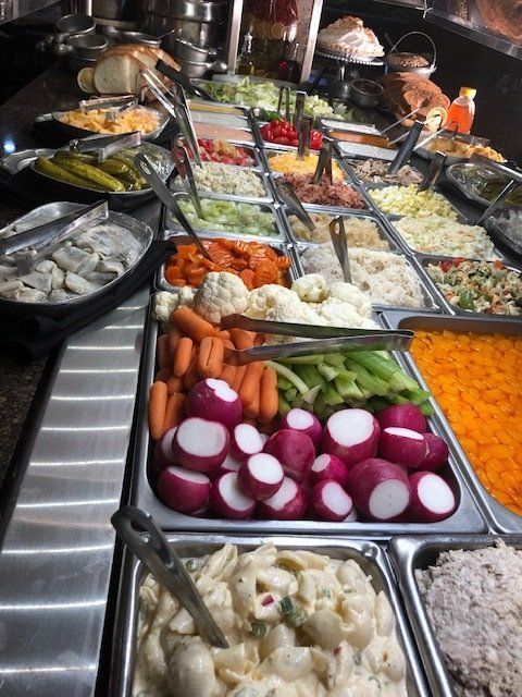 Buffet counter with assorted salads, vegetables, and seafood in metal trays and tongs