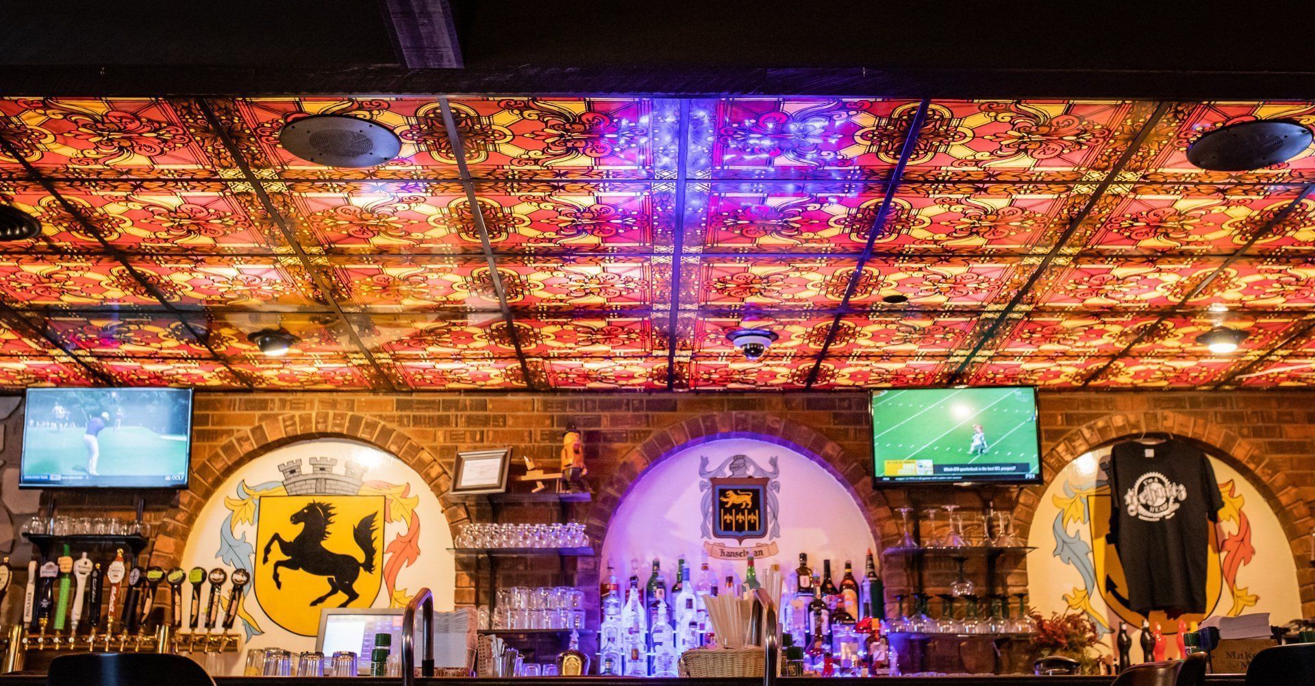 Colorful bar interior with illuminated ceiling, wall-mounted TVs, arched decor, and bottles lined along the counter