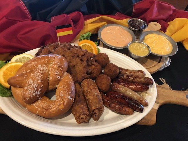Pretzel, sausage, croquettes, and dipping sauces on a plate, with German flags in the background