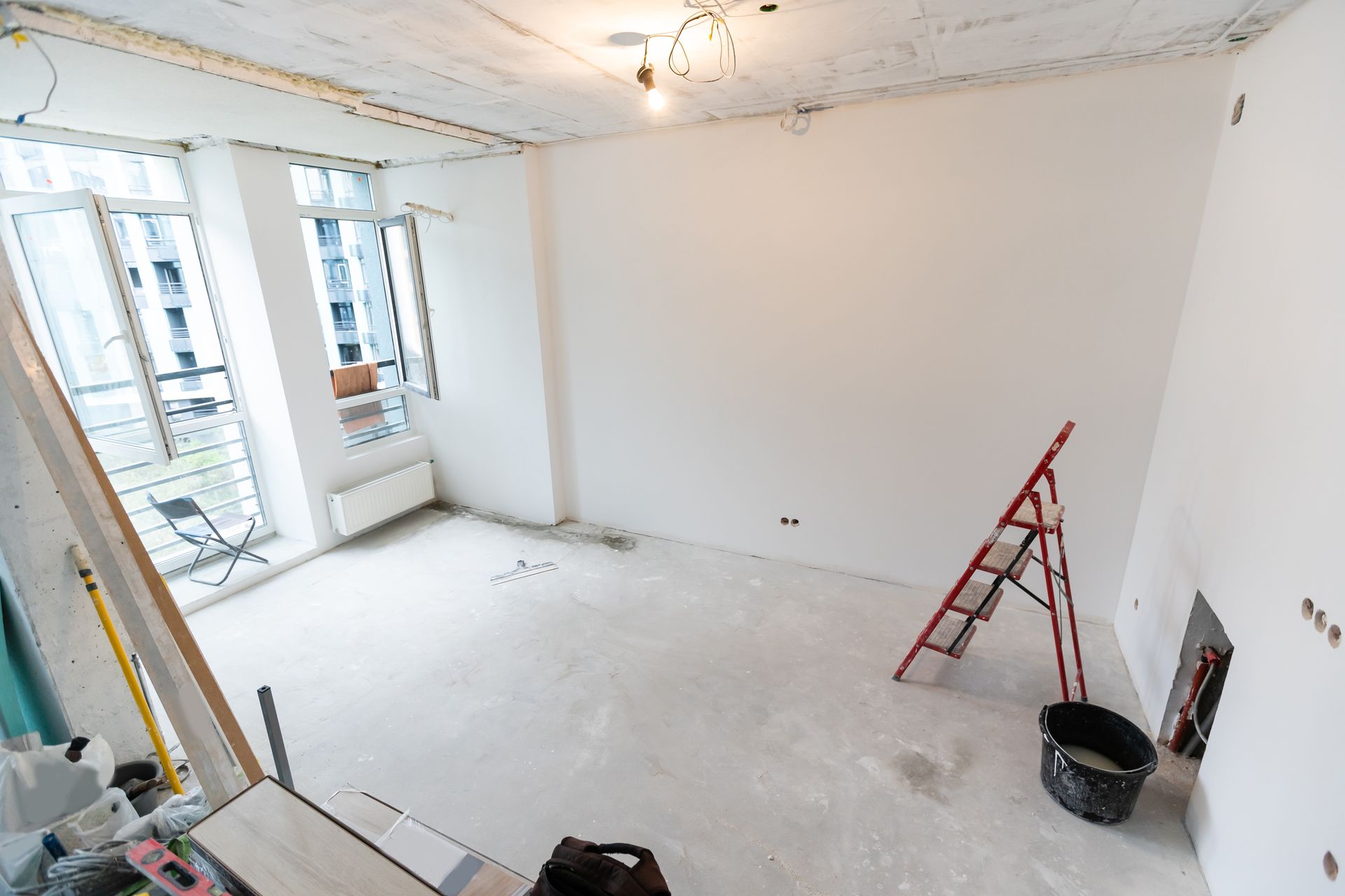 Empty room under renovation with white walls, exposed ceiling, red ladder, and paint bucket.