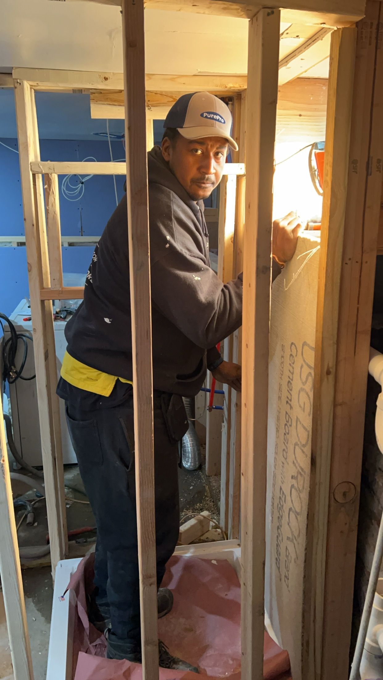 Worker in a cap and dark jacket standing inside a partially framed room during construction.