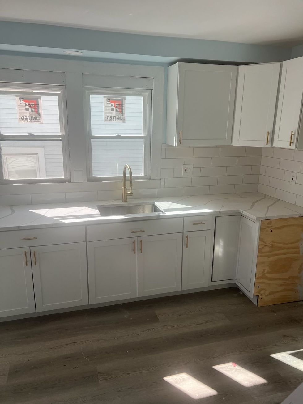 Bright empty kitchen with white cabinets, window over sink, and sunlight on dark floor.