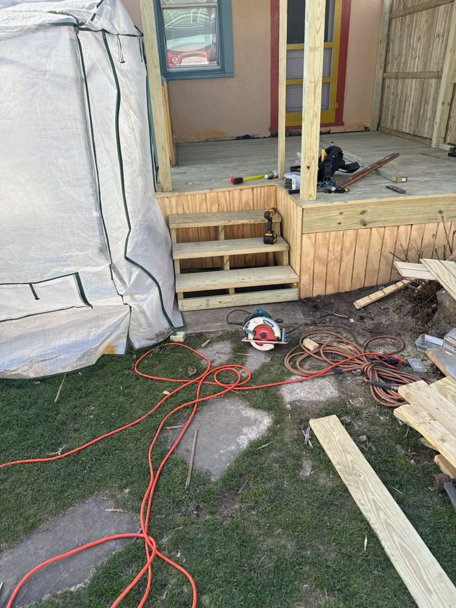 Construction debris and tools scattered on a grassy yard beside a small wooden deck and house entry.