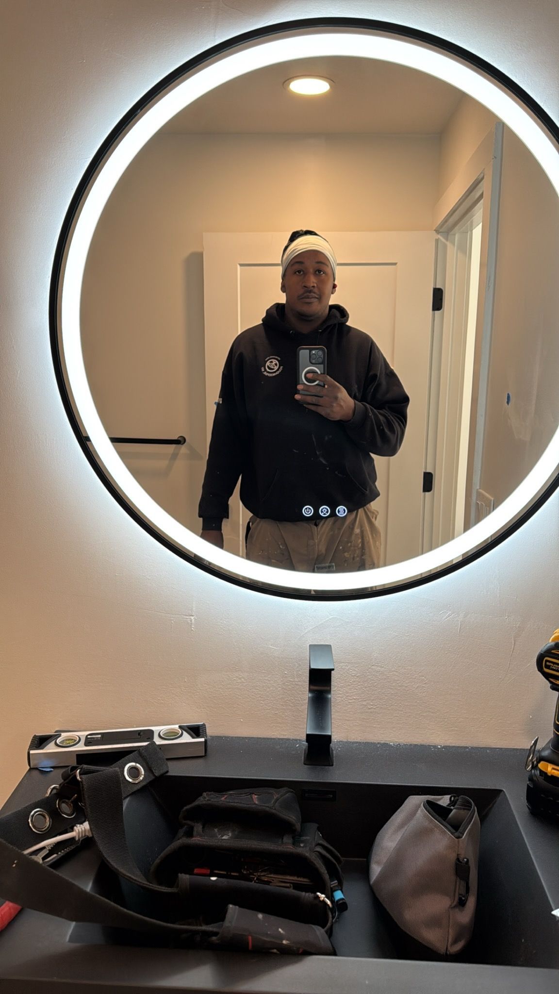 Person taking a mirror selfie in a bathroom, with a round backlit mirror and black countertop.