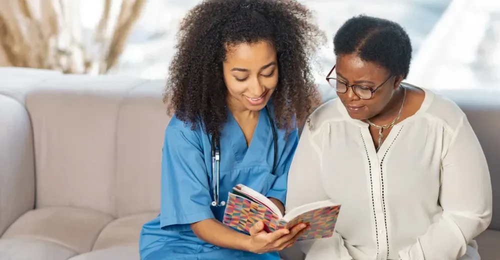 Two people sit on a couch, reading a book together and smiling.
