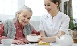 Caregiver serving tea to an elderly woman at a table, with cups, teapot, and pastries.