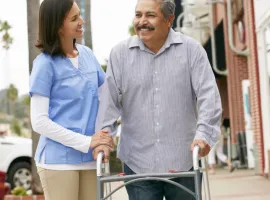 Caregiver helping a man walk with a walker on a sidewalk outside a building