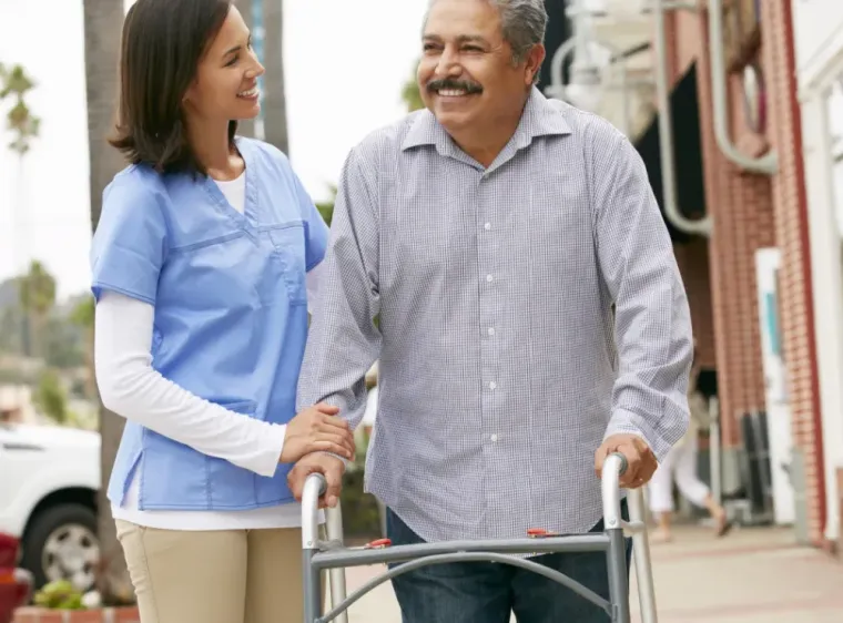 Two people smiling while walking with a walker on a sidewalk, one in blue scrubs assisting.