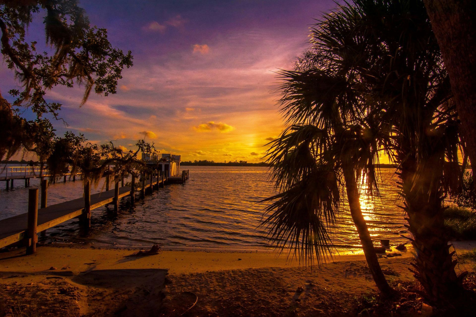 Sunset over a calm lake. A wooden pier extends into the water. Palm trees frame the colorful sky.