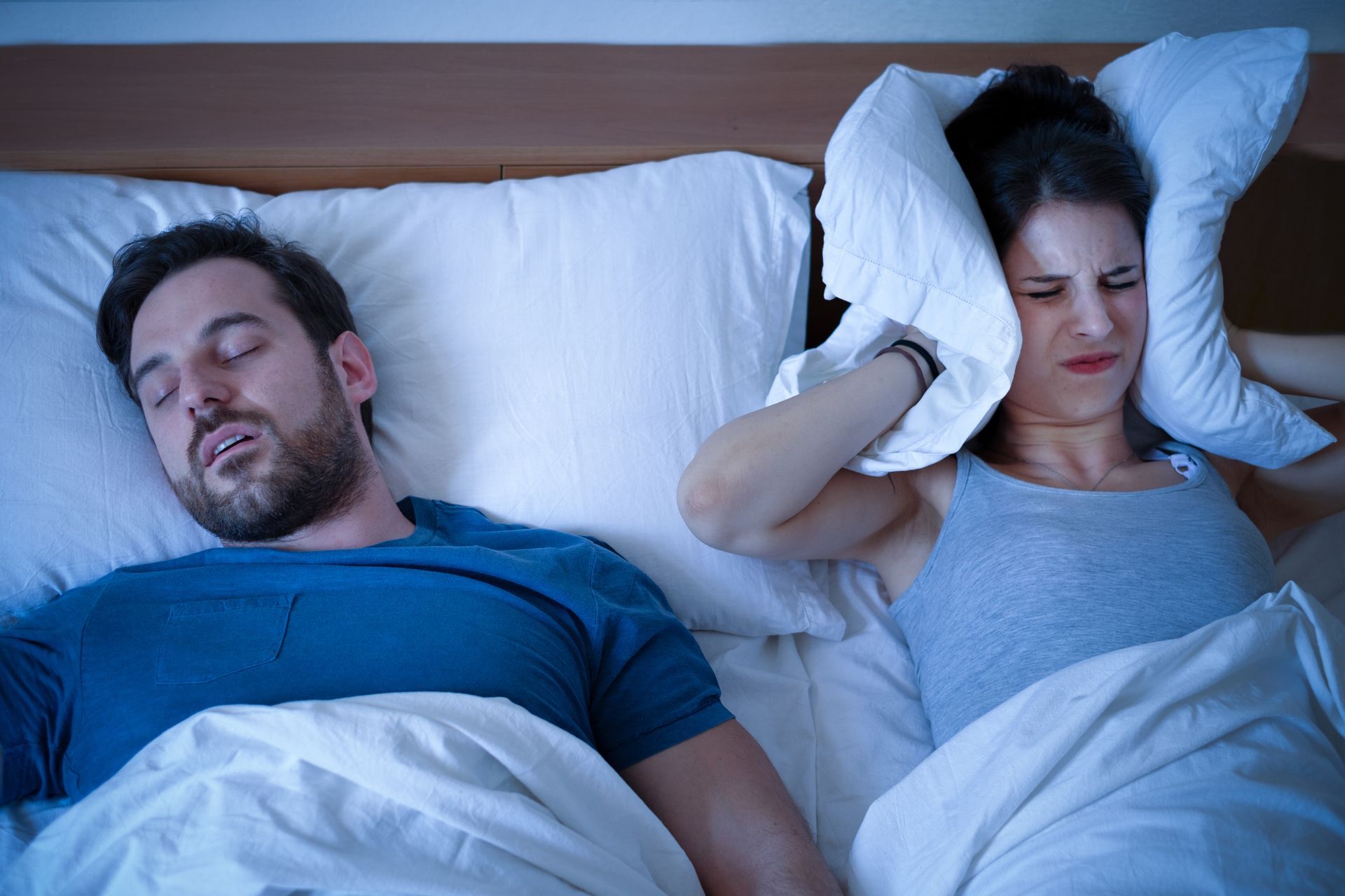 A man is snoring while a woman covers her ears with pillows.