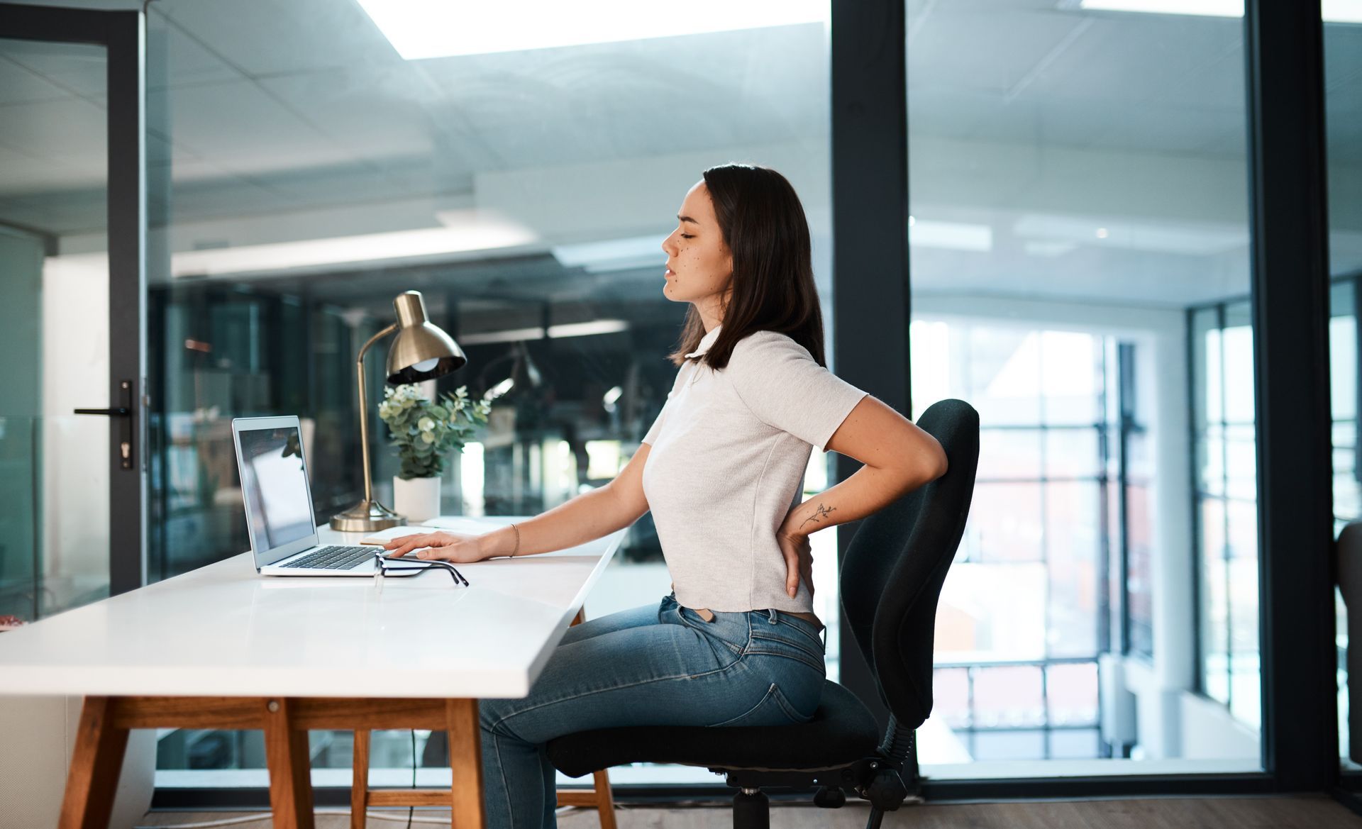 A woman is sitting at a desk with a laptop and holding her back.