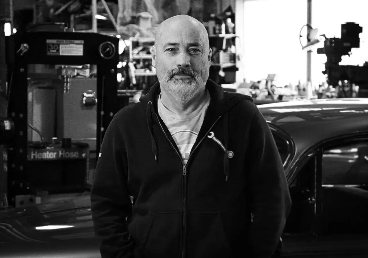 Man standing in a workshop beside machinery, with a car in the background. Man standing in a workshop beside machinery, with a car in the background.