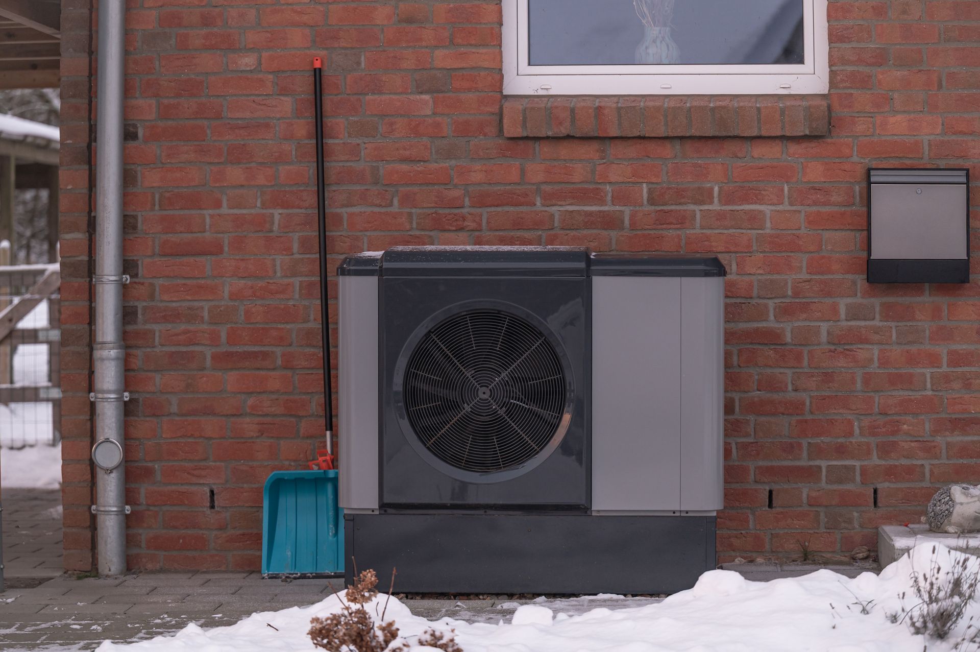Large black HVAC unit beside a brick house, with snow on the ground and a blue bin nearby