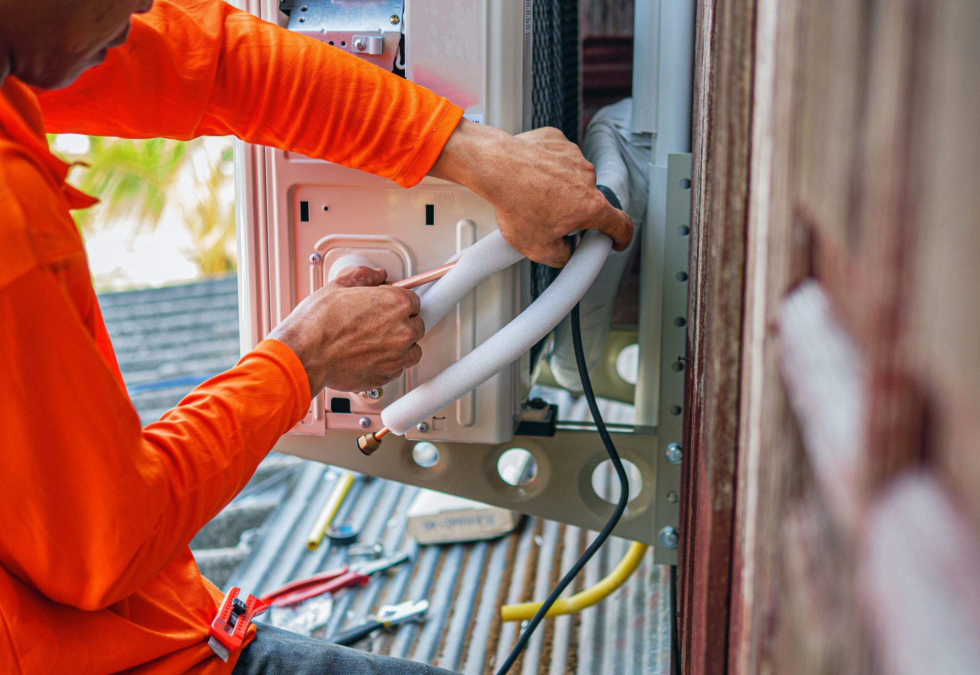 Technician in orange shirt wiring an outdoor HVAC unit on a building wall