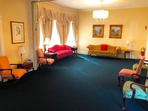 A room with blue carpet, couches, and chairs. Pink and yellow decor, large windows.