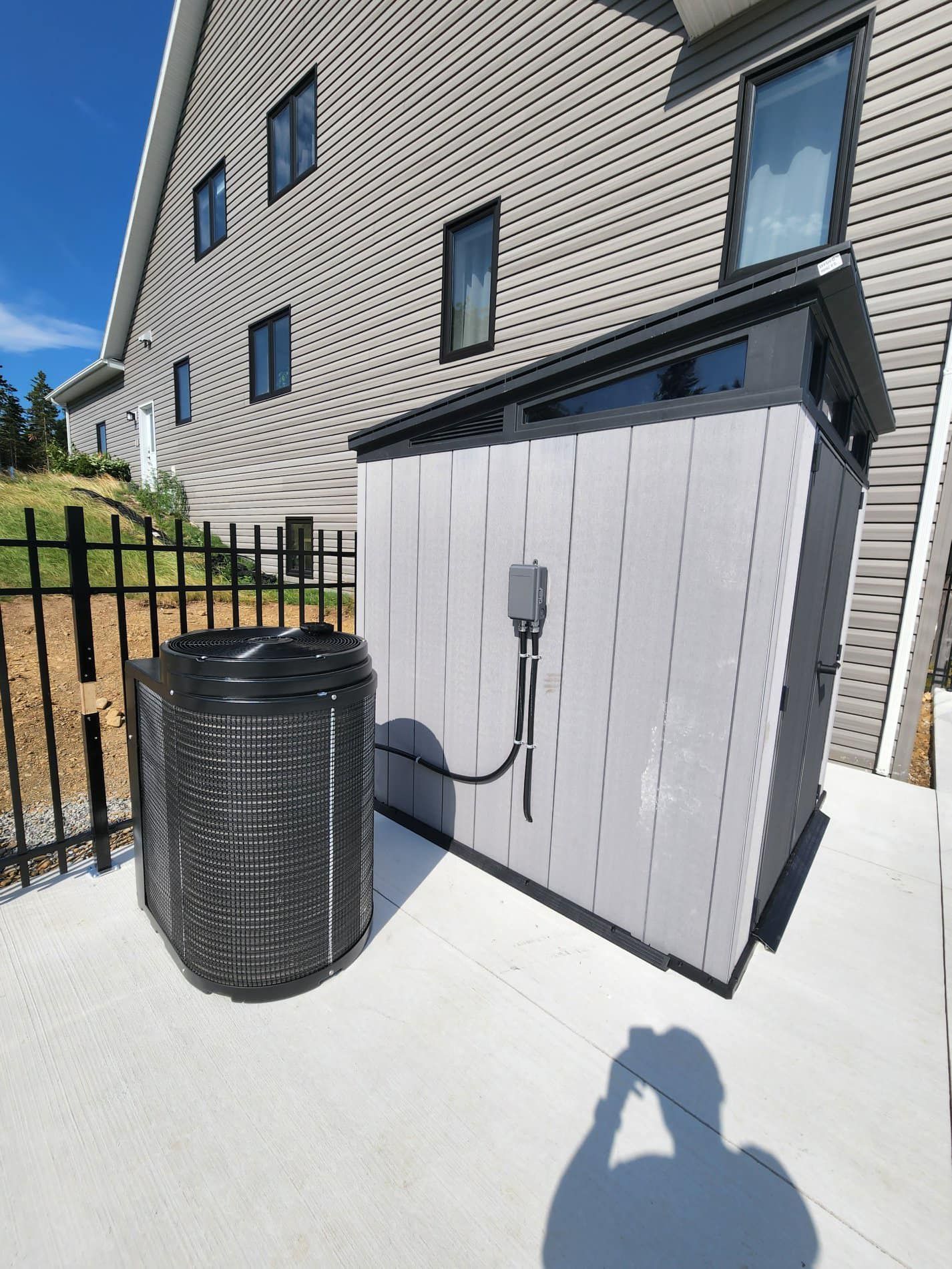 A black air conditioner is sitting next to a gray shed in front of a house.