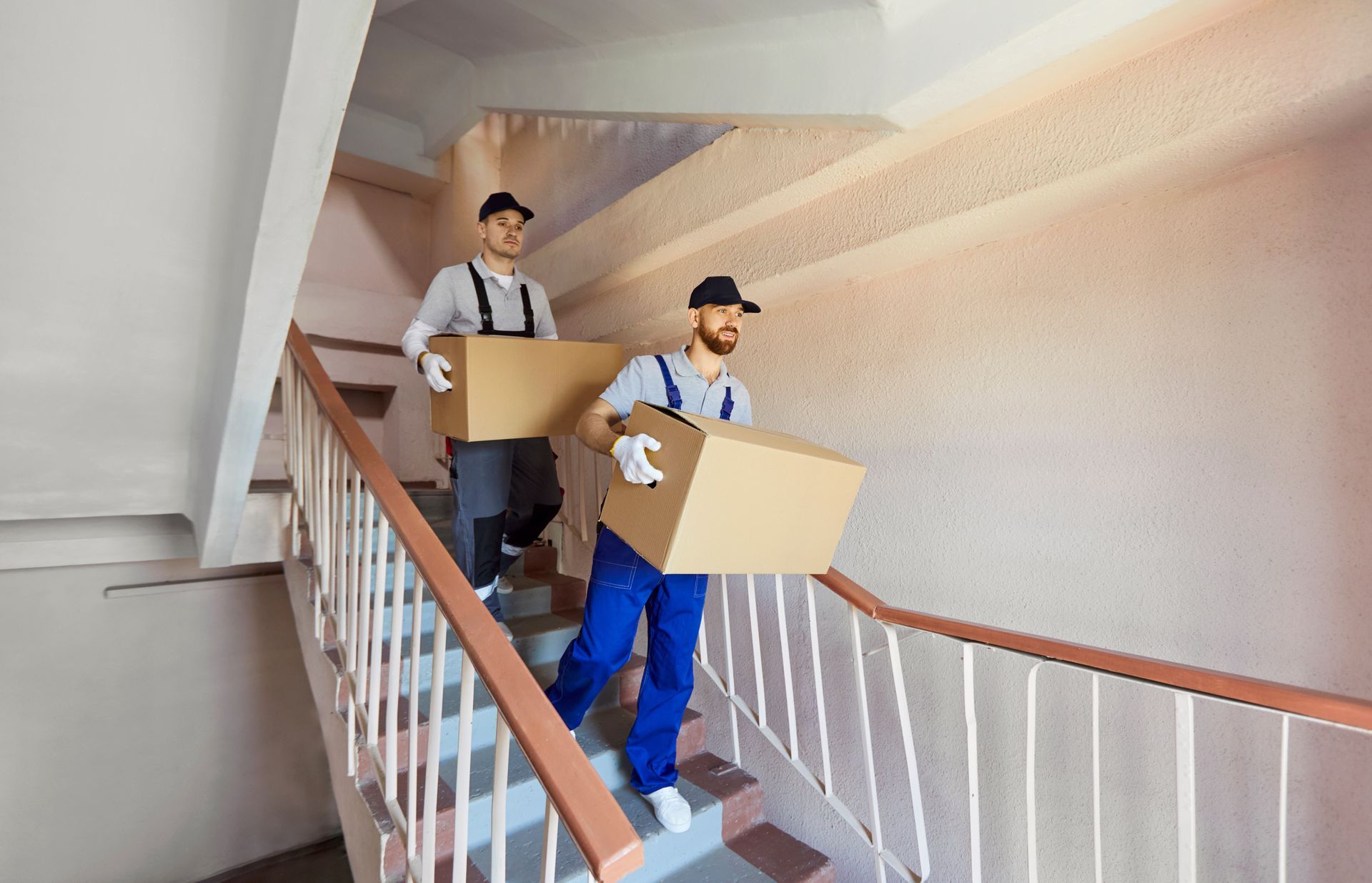 Two movers carrying boxes down a stairwell, one in blue overalls and one in a striped shirt