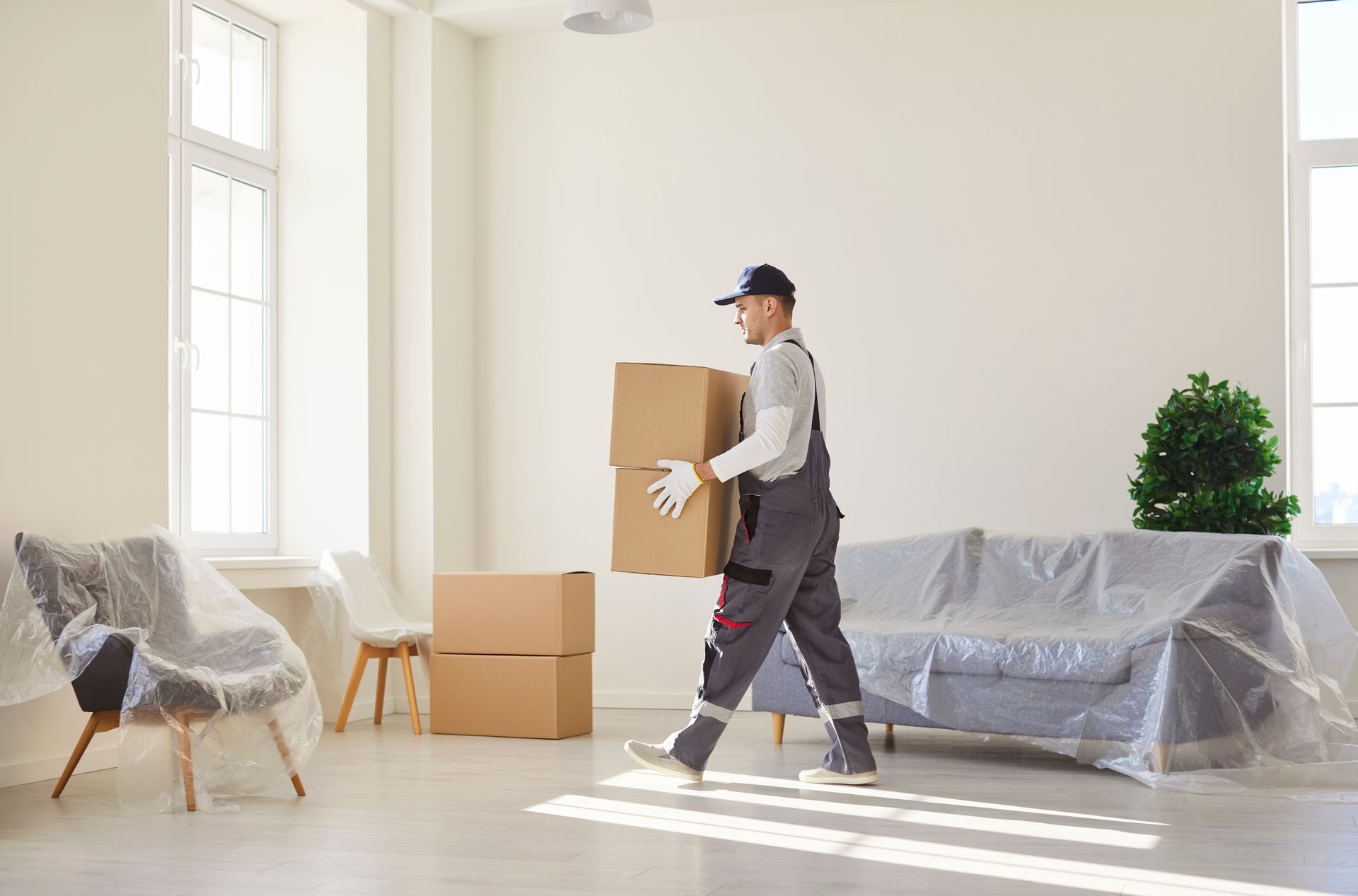 Mover carrying a box in a bright living room with furniture covered in plastic and stacked moving boxes.