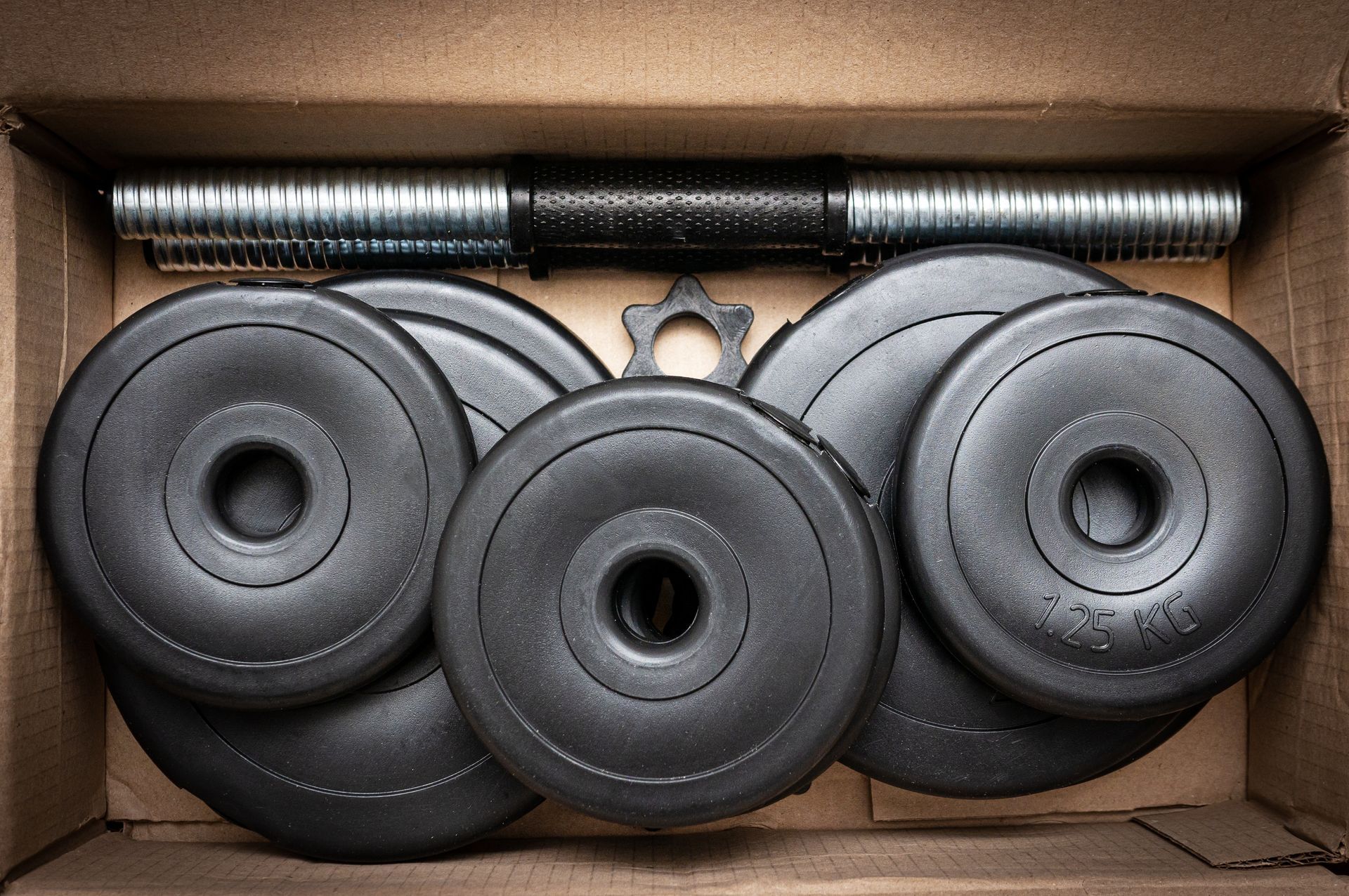 Box of black rubber weight plates and a barbell sleeve stacked in a cardboard box